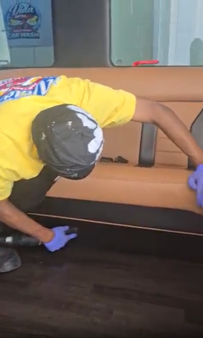 A Vida Detail technician hand‑cleaning the leather door panel and wood trim of a vehicle interior.